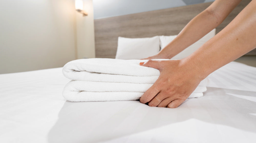 Close-up of hands putting stack of fresh white bath towels on the bed sheet