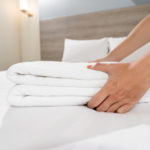 Close-up of hands putting stack of fresh white bath towels on the bed sheet | How Often Should You Replace Your Airbnb Linens And Towels | how often should you replace your towels | how often to replace towels | Featured