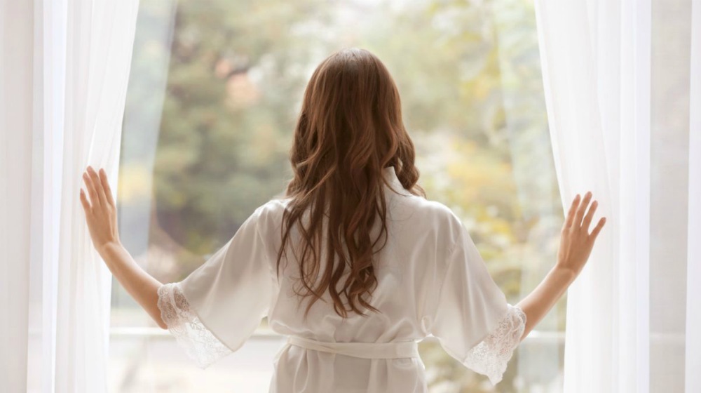 woman in a silk robe standing near window | Satin and Silk Robes: How To Wear and Care For Them | silk robe | long silk robe