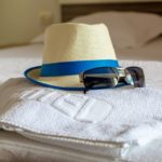 hat towel and sunglasses on bed | Advantages Of Using Personalized Beach Towels To Promote Business | beach towels | custom beach towels | Featured