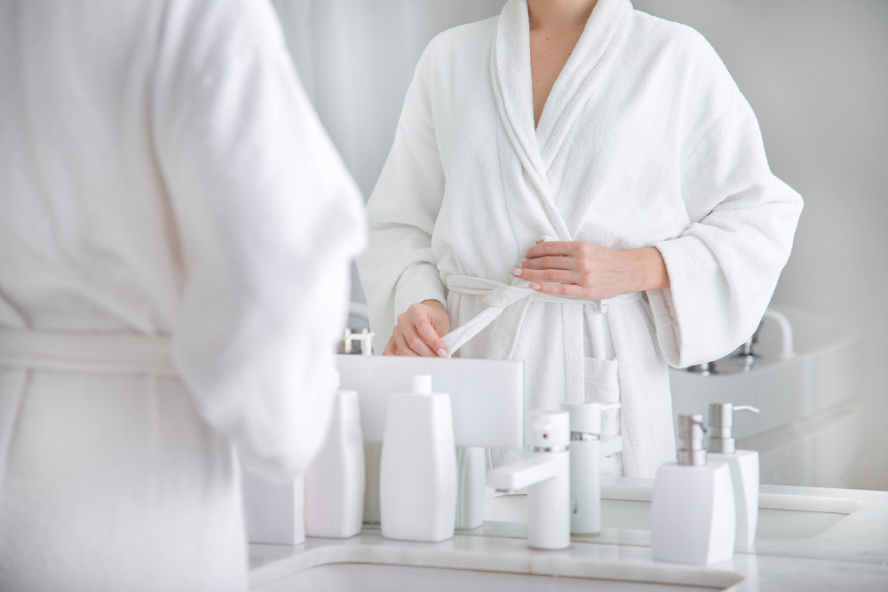 Close up of body of young woman tying up belt of white bathrobe in front of the mirror | Airbnb Bathroom Essentials To Stock For Your Guests | airbnb supplies